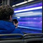 Passager i en togkupé om aftenen set skråt bagfra taler til sin telefon, mens byens neonskær glider forbi vinduet