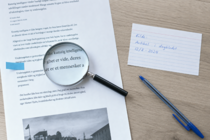 Skrivebord set lige oppefra med en printet artikelside, hvor ét afsnit er markeret med et blåt faneblad, et forstørrelsesglas over de trykte linjer og et håndskrevet kildekort ved siden af
