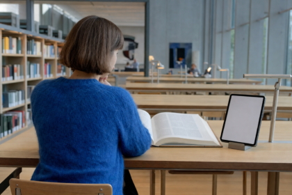 Person set skråt bagfra i halvtotal ved et langt egetræsbord i en lys skandinavisk læsesal, med et opslået opslagsværk til venstre og en opretstående tablet til højre i blødt morgenlys fra siden