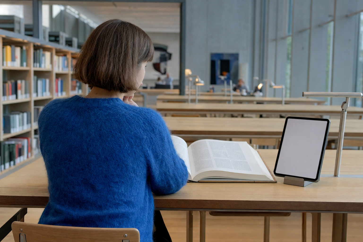 Person set skråt bagfra i halvtotal ved et langt egetræsbord i en lys skandinavisk læsesal, med et opslået opslagsværk til venstre og en opretstående tablet til højre i blødt morgenlys fra siden