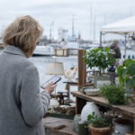 Person i grå frakke set skråt bagfra ved en loppemarkedsbod på en dansk havnekaj kigger på en mobil med en ny notifikation, overskyet dagslys og grønne planter på boden