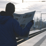 Pendler på en perron holder en avis foldet helt ud, så den dækker alt under den, set skråt bagfra i modlys