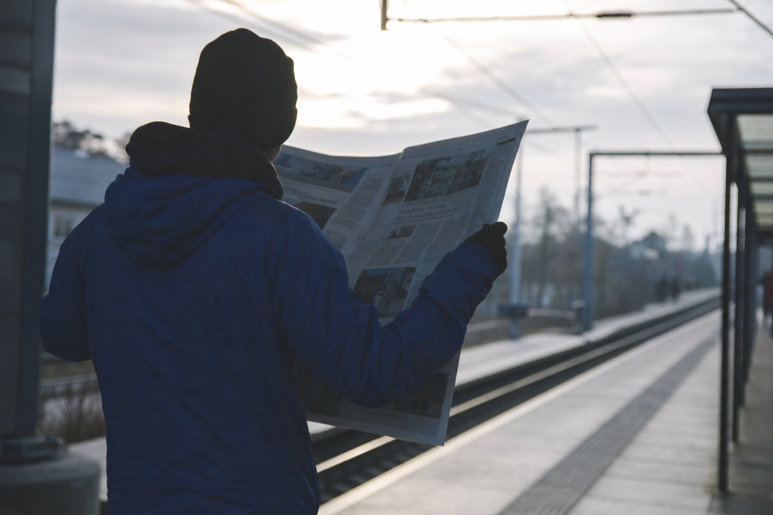 Pendler på en perron holder en avis foldet helt ud, så den dækker alt under den, set skråt bagfra i modlys