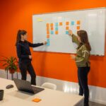 To kvinder står foran en whiteboard med noter i et lyst kontor og drøfter en plan, orange accent i ellers neutral scene