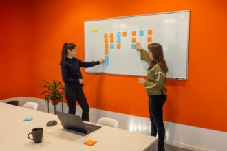 To kvinder står foran en whiteboard med noter i et lyst kontor og drøfter en plan, orange accent i ellers neutral scene