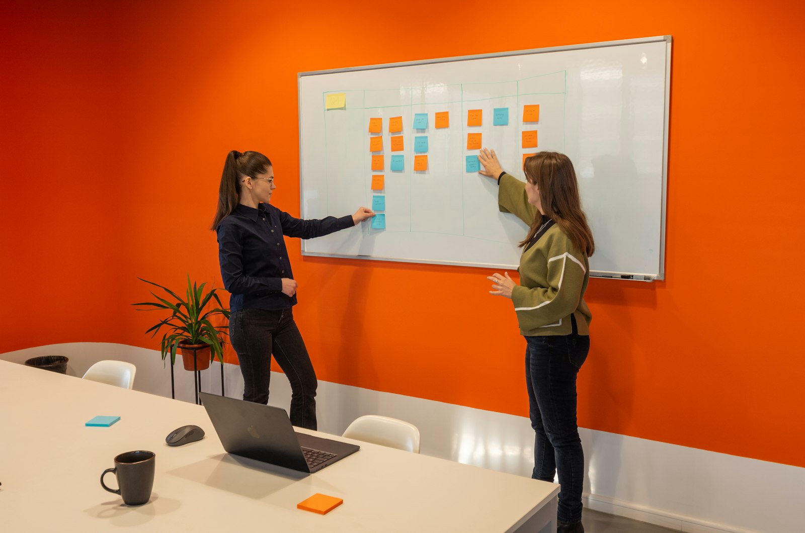 To kvinder står foran en whiteboard med noter i et lyst kontor og drøfter en plan, orange accent i ellers neutral scene