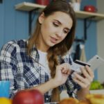 Kvinde ved et bord med smartphone i den ene hånd og betalingskort i den anden, midt i et onlinekøb