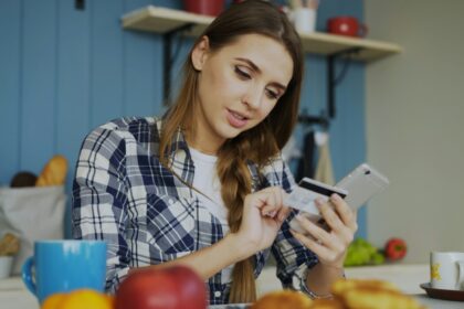 Kvinde ved et bord med smartphone i den ene hånd og betalingskort i den anden, midt i et onlinekøb