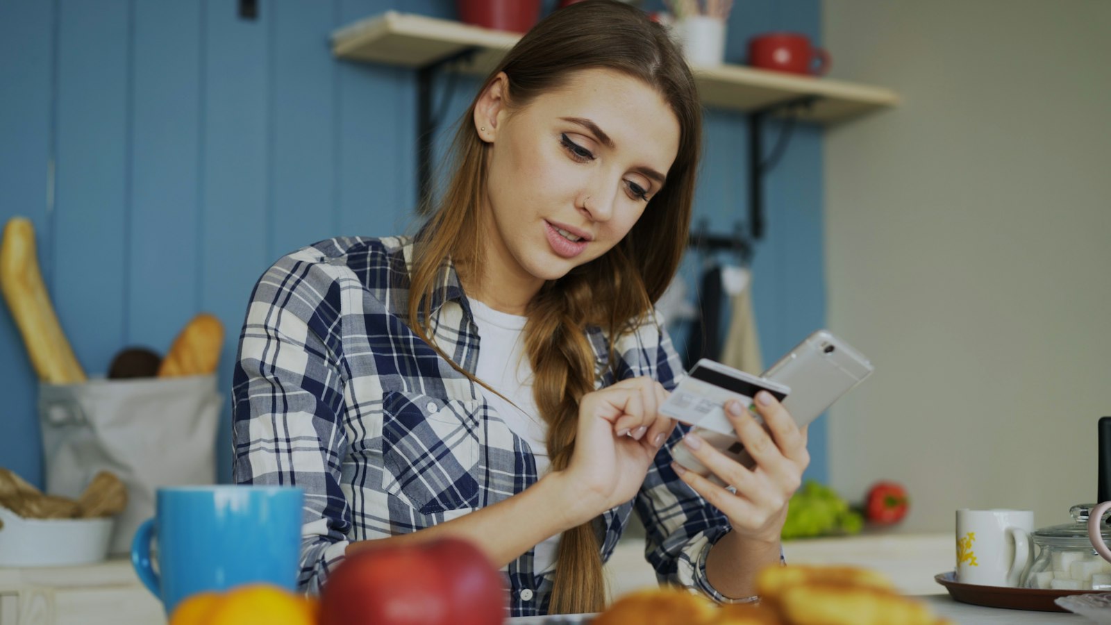 Kvinde ved et bord med smartphone i den ene hånd og betalingskort i den anden, midt i et onlinekøb