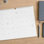 Skrivebord set direkte fra oven i neutralt dagslys med en papirkalender, hvor tre datoer er markeret med blå ring, ved siden af notesbog, pen og telefon