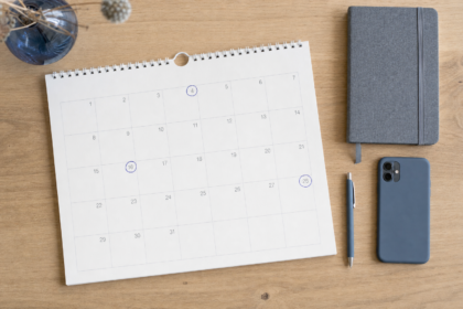 Skrivebord set direkte fra oven i neutralt dagslys med en papirkalender, hvor tre datoer er markeret med blå ring, ved siden af notesbog, pen og telefon