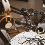Nærbillede af en grå og sort kondensatormikrofon i et mørkt podcaststudie, varmt lampelys mod sort baggrund