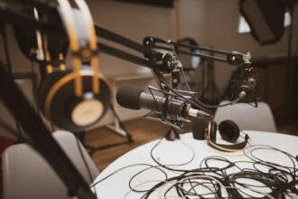 Nærbillede af en grå og sort kondensatormikrofon i et mørkt podcaststudie, varmt lampelys mod sort baggrund