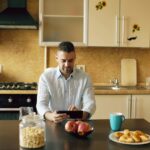 Mand ved køkkenbord om morgenen ser på en tablet mellem morgenmad og kaffe